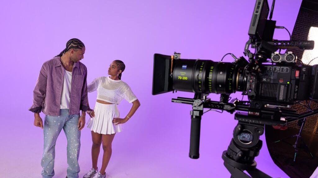 A professional camera equipped with a ZEISS lens captures two people posing on a purple-lit studio set.