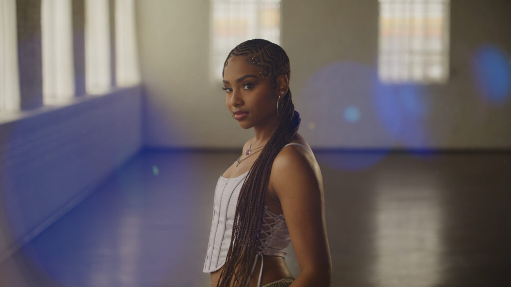 A woman with long braided hair stands in a softly lit, spacious room with large windows and lens flares, captured with cinematic clarity.