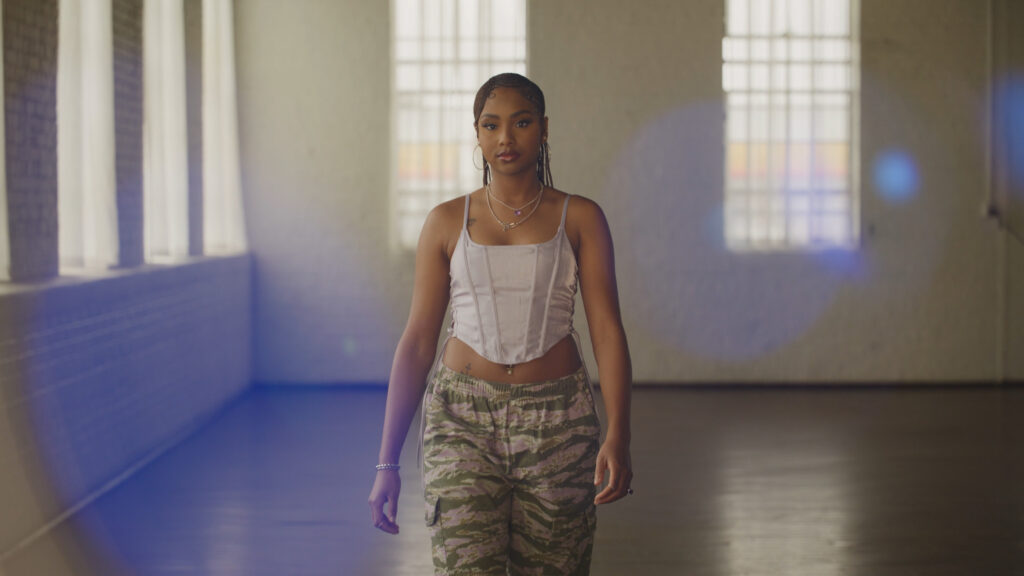 A woman in a white corset top and camouflage pants walks confidently in a sunlit indoor space with lens flare effects, showcasing ZEISS lens clarity.
