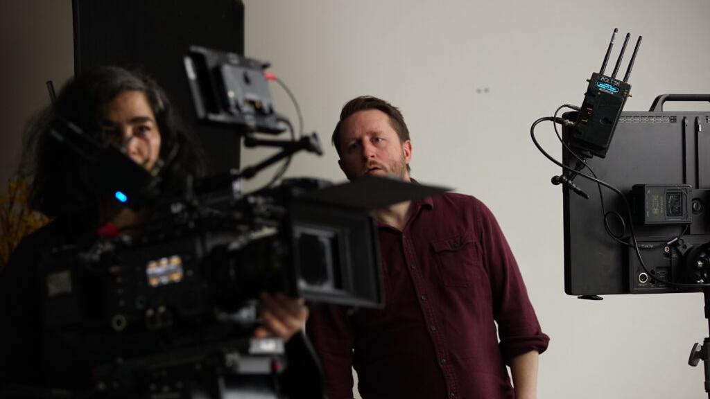 Two people are working with professional film production equipment, including a camera and monitor, in a studio setting featuring ZEISS lenses.