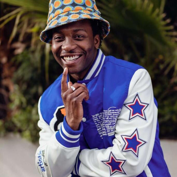 A smiling man wearing a colorful polka-dot bucket hat and a blue varsity jacket with star patches poses outdoors in front of green palm leaves.