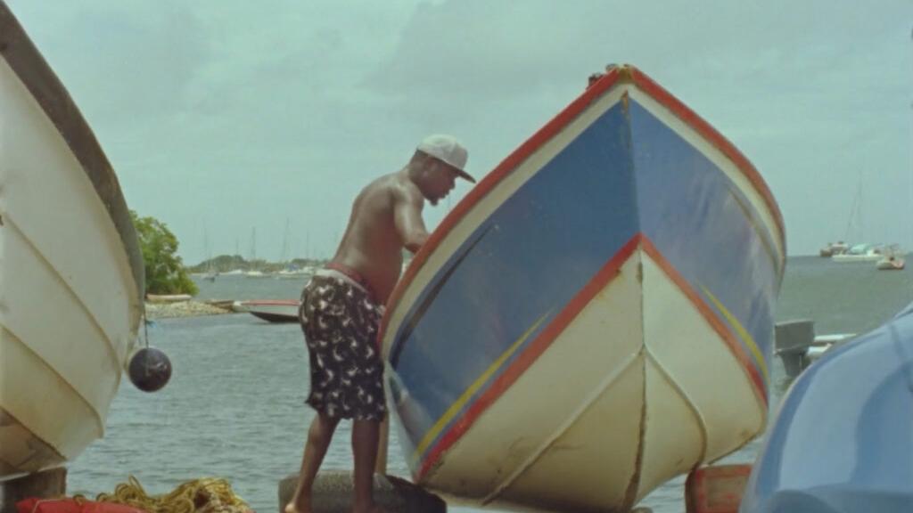 A man stands on a dock next to a colorful fishing boat with the ocean and other boats visible in the background.