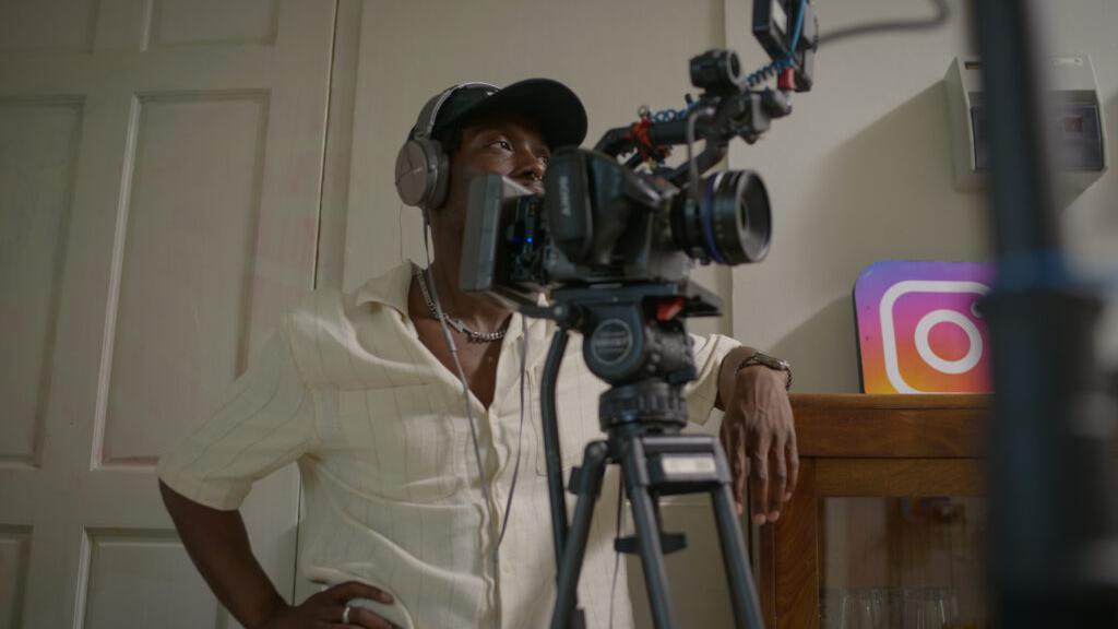 A man wearing headphones stands behind a professional video camera setup equipped with accessories, with a visible ZEISS lens.