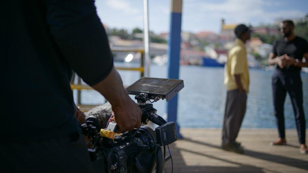 A professional camera setup equipped with a ZEISS lens captures two people having a conversation on a dock by the water.