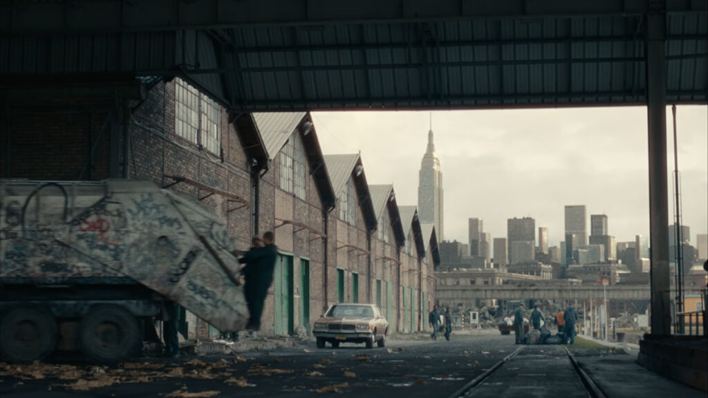 A man wearing a gray blazer, black shirt, chain necklace, and ripped jeans sits in a modern, dimly lit room with large windows and indoor plants, captured with ZEISS lens clarity.