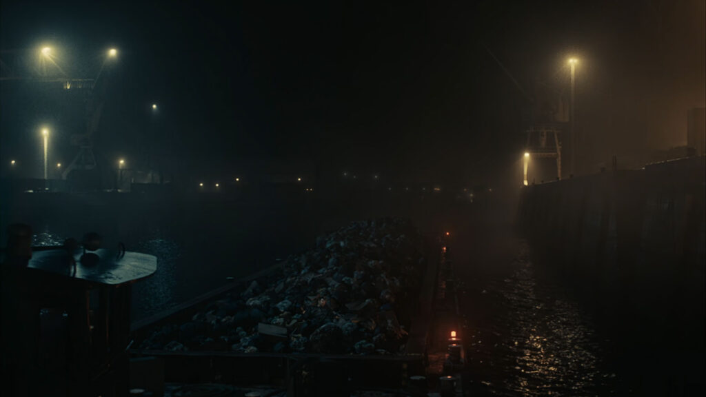 A man wearing a gray blazer, black shirt, chain necklace, and ripped jeans sits in a modern, dimly lit room with large windows and indoor plants, captured with ZEISS lens clarity.