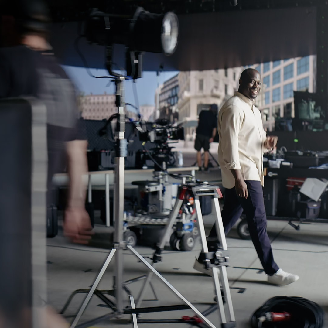 A man walks through a professional film set equipped with lighting, cameras, and other production gear, with a cityscape backdrop and ZEISS camera equipment in use.