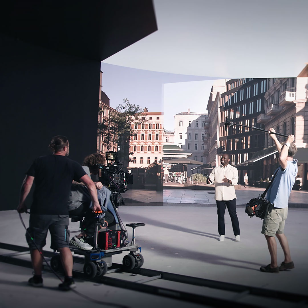 A film crew operating a professional camera and crane on an outdoor set, capturing a presenter against a virtual cityscape backdrop using ZEISS technology.