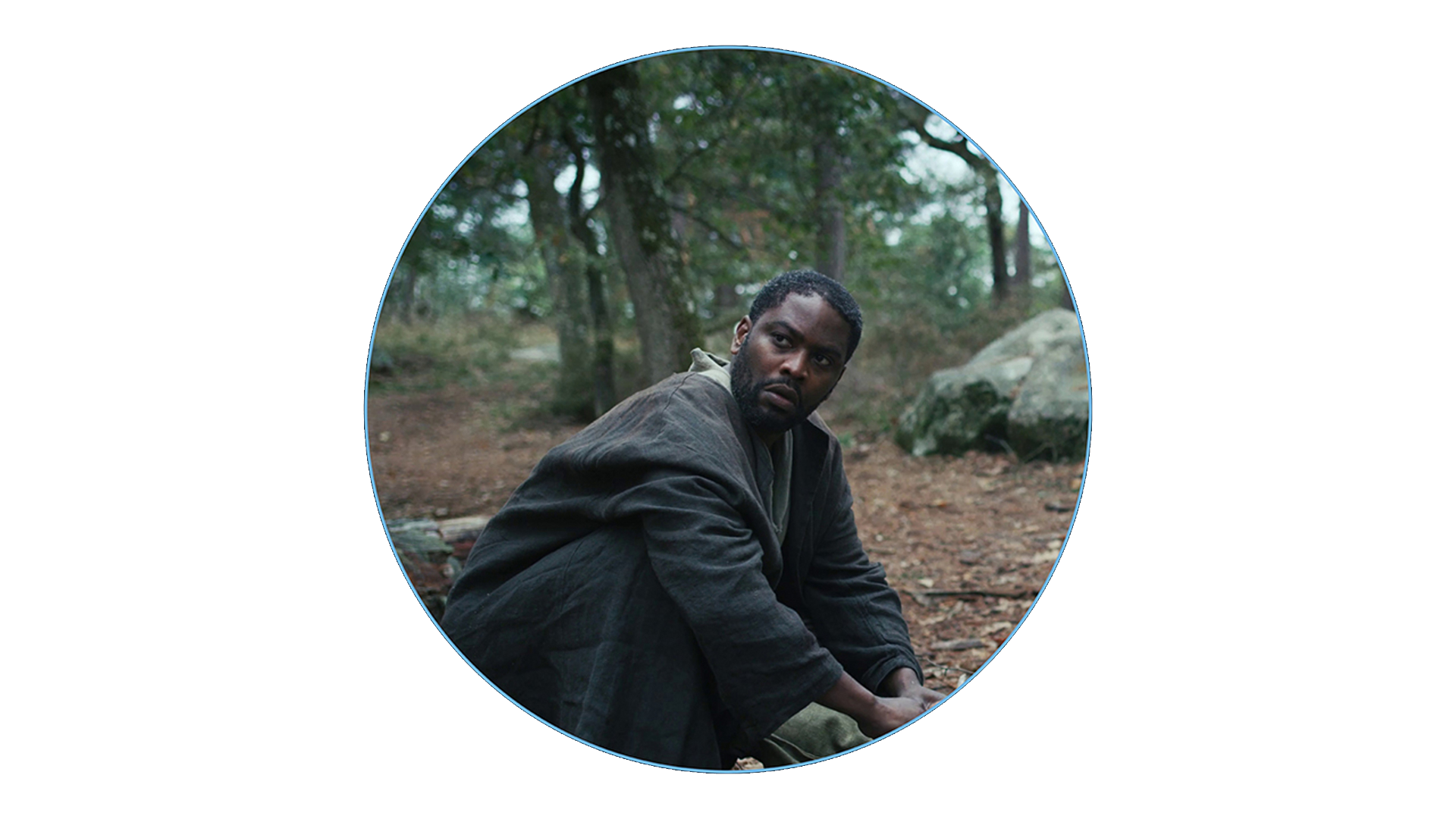 A man in dark clothing is sitting on the forest floor surrounded by trees and rocks, framed within a circular ZEISS lens view.