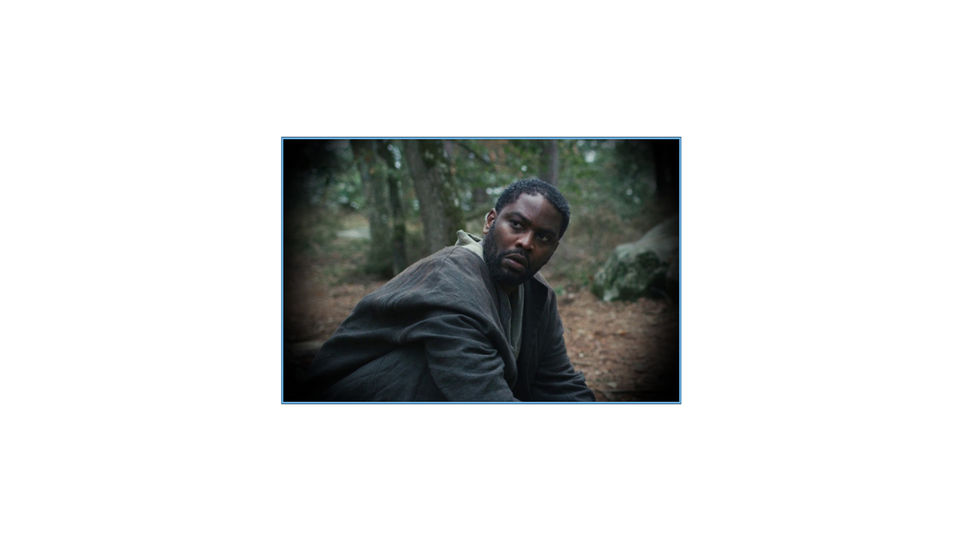A man is sitting in a forest clearing, seen through the circular view of a ZEISS lens.