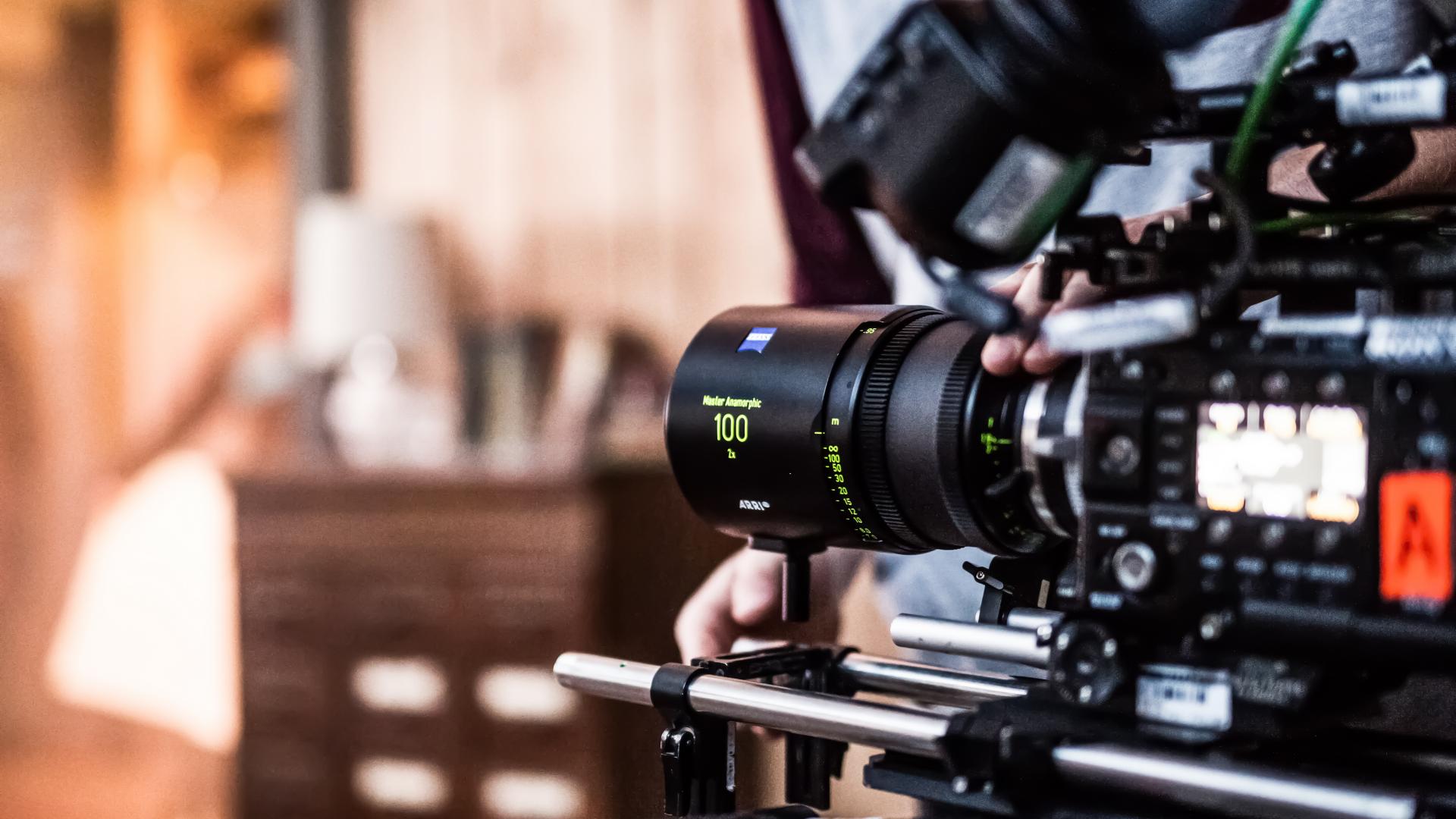 A professional cinema camera is equipped with a ZEISS Supreme Prime 100mm lens being adjusted by a crew member on set.
