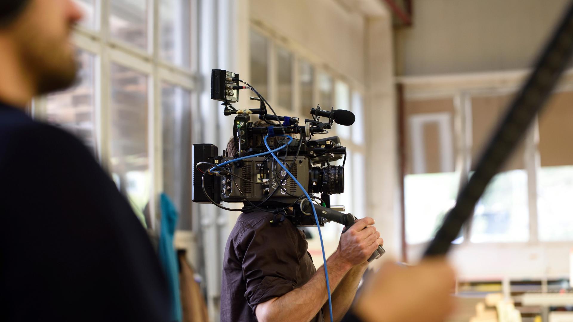A camera operator is filming with professional video equipment, including a ZEISS lens, in a brightly lit indoor setting.