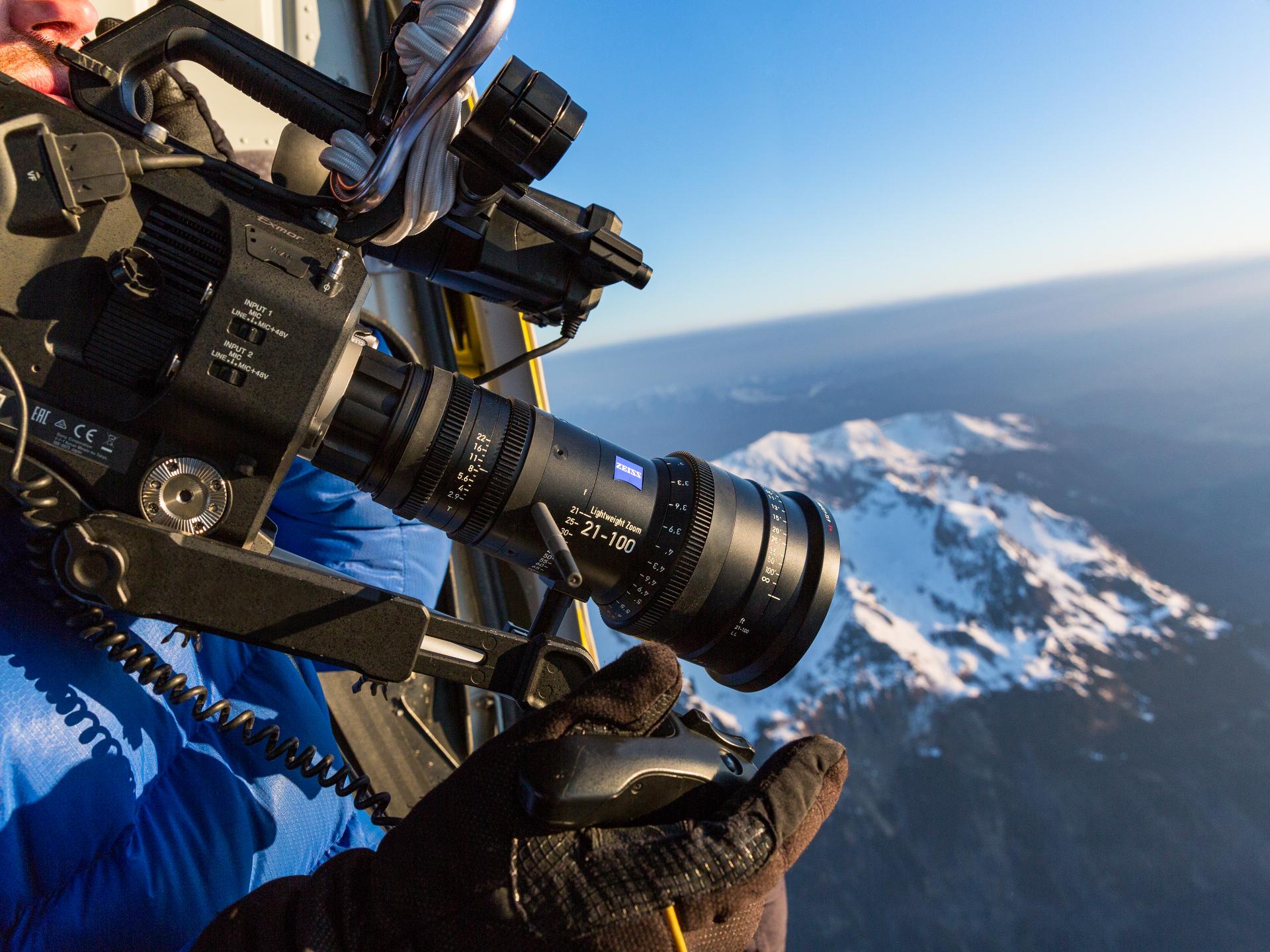 A professional camera equipped with a ZEISS lens is being operated from a helicopter to film a snow-covered mountain landscape.