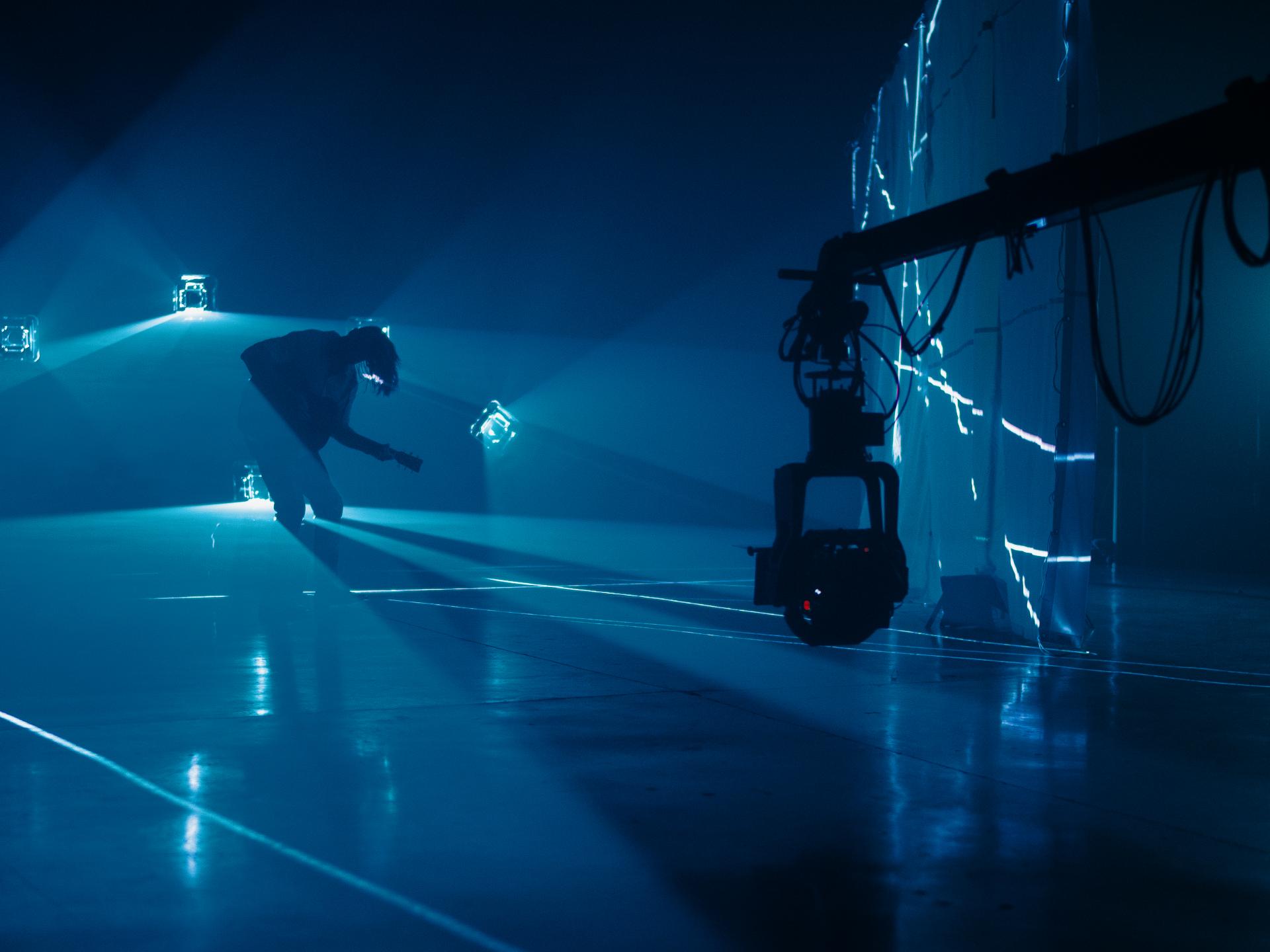 A person is performing on a dark stage illuminated by blue lights, with a large camera rig in the foreground and light patterns projected on the background wall, showcasing ZEISS imaging technology.