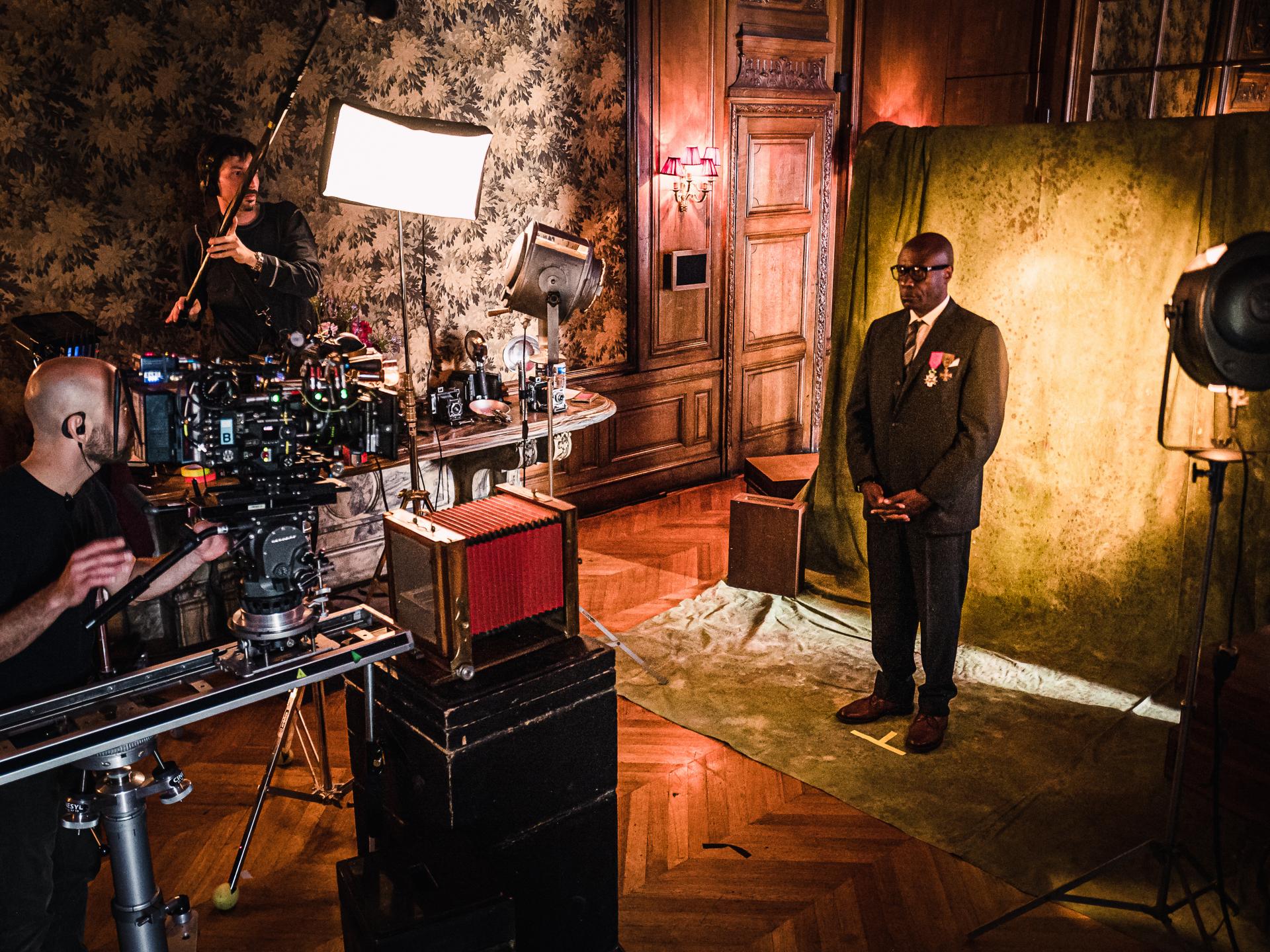 A film crew captures a formally dressed man standing in front of a textured backdrop in an ornate room, using professional ZEISS camera equipment.