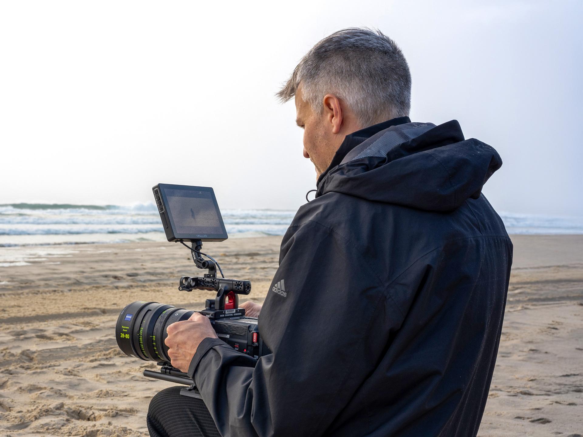 A man operating a professional video camera with a ZEISS lens on a beach.