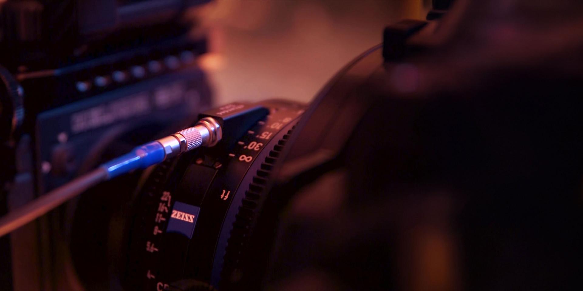 Close-up view of a professional camera lens with ZEISS branding and connected cable in a low-light setting.