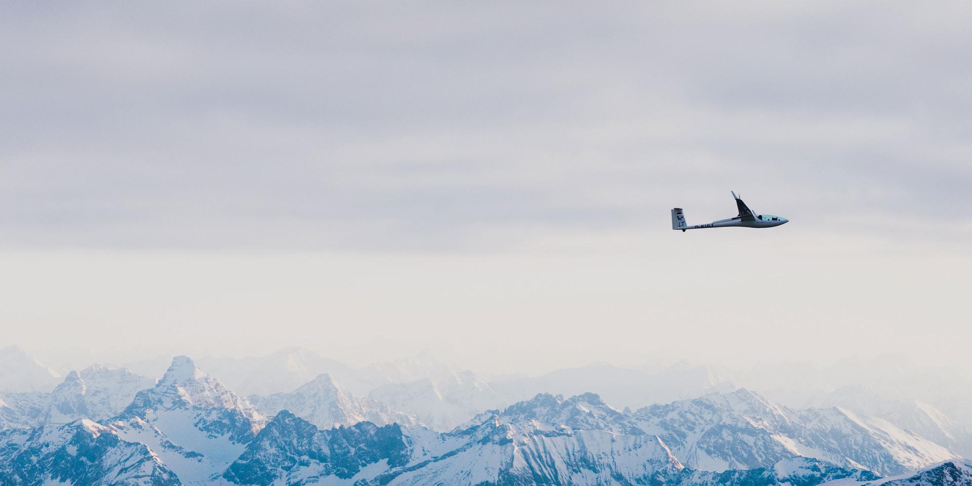 A glider soars above a vast range of snow-covered mountains under a cloudy sky.