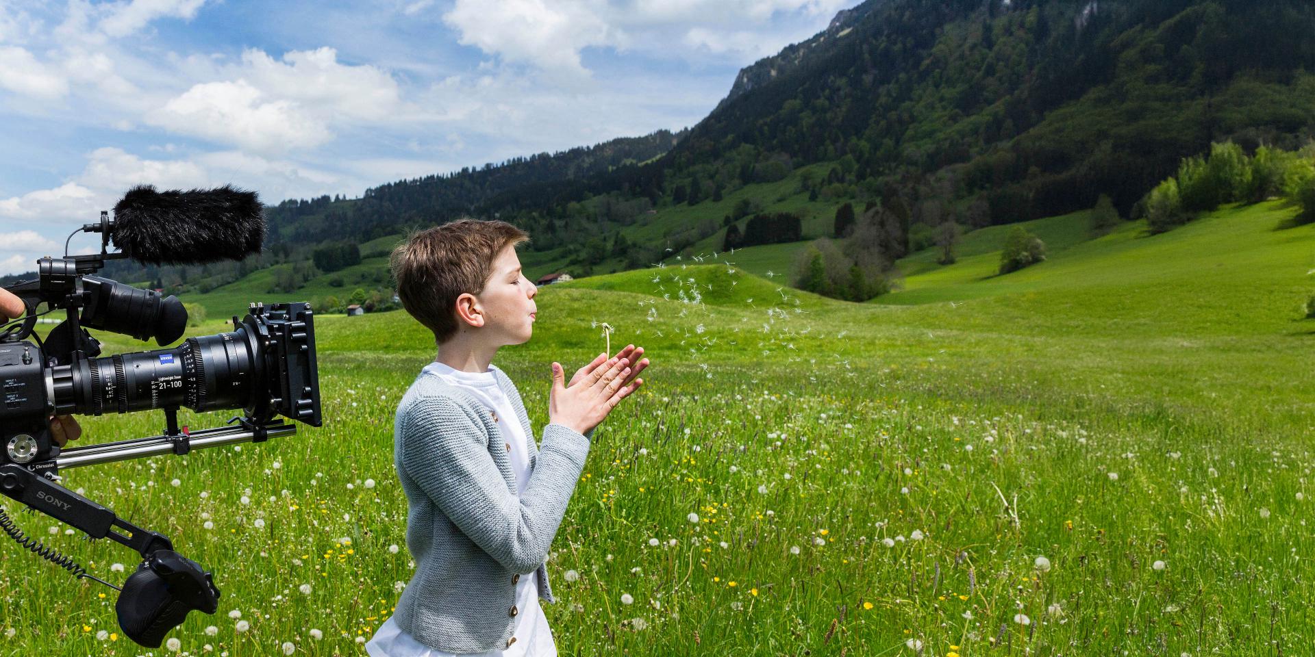 A professional camera films a child standing in a green meadow with mountains in the background under a partly cloudy sky.