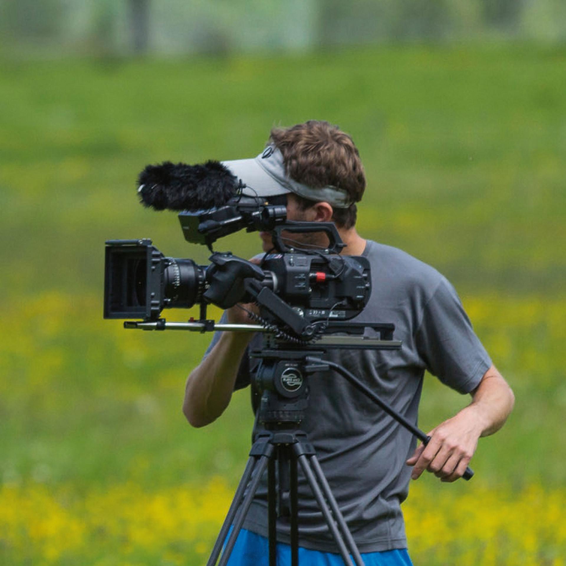 A man is operating a professional video camera with a mounted microphone on a tripod in an outdoor field.