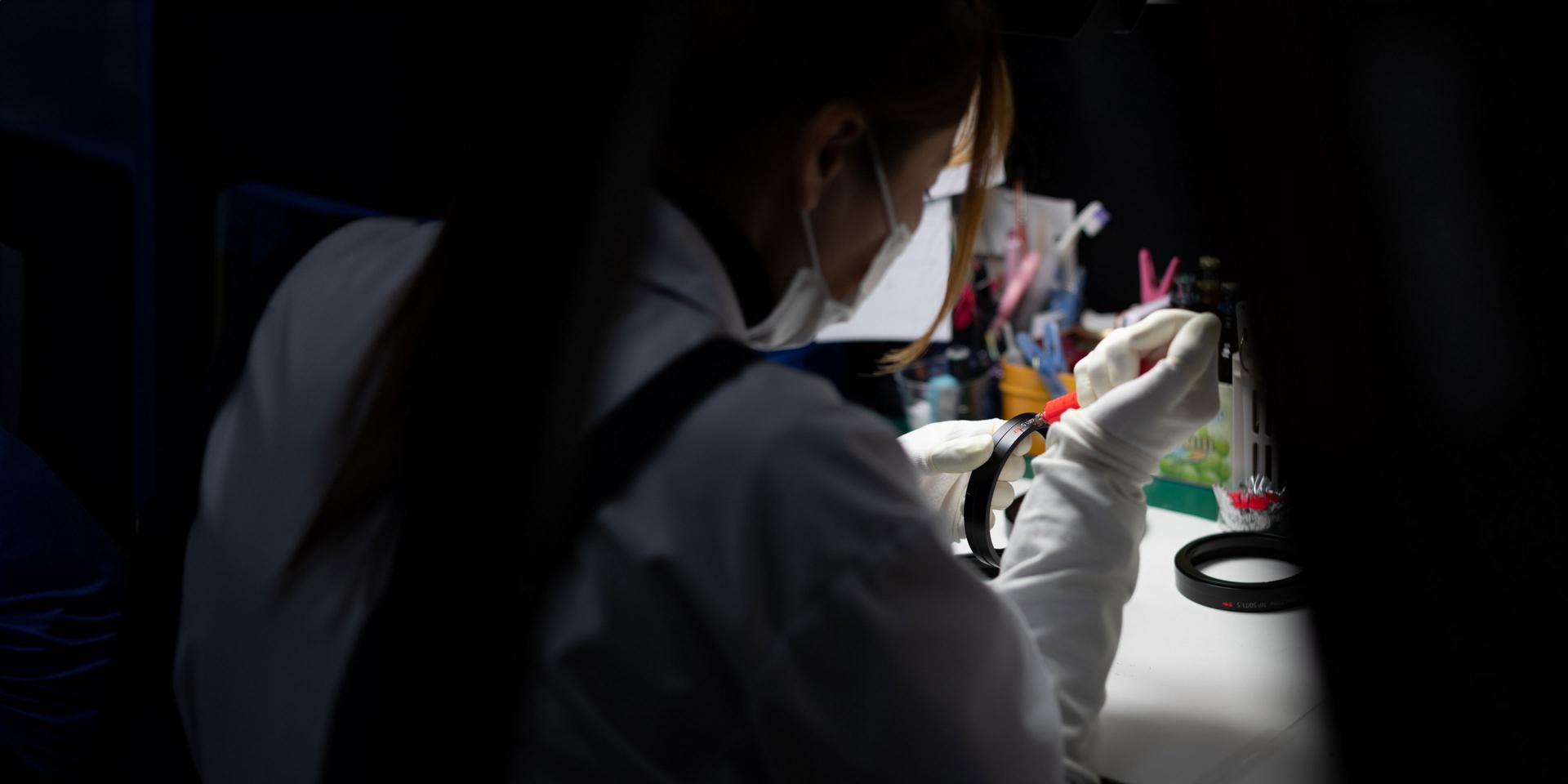 A technician wearing a lab coat and gloves works with precision tools under focused lighting at a ZEISS workstation.