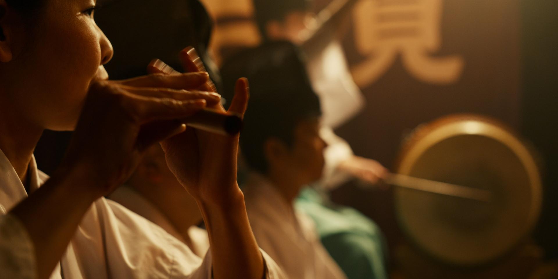 Musicians in traditional attire play Japanese instruments, including flutes and a drum, in a softly lit setting.