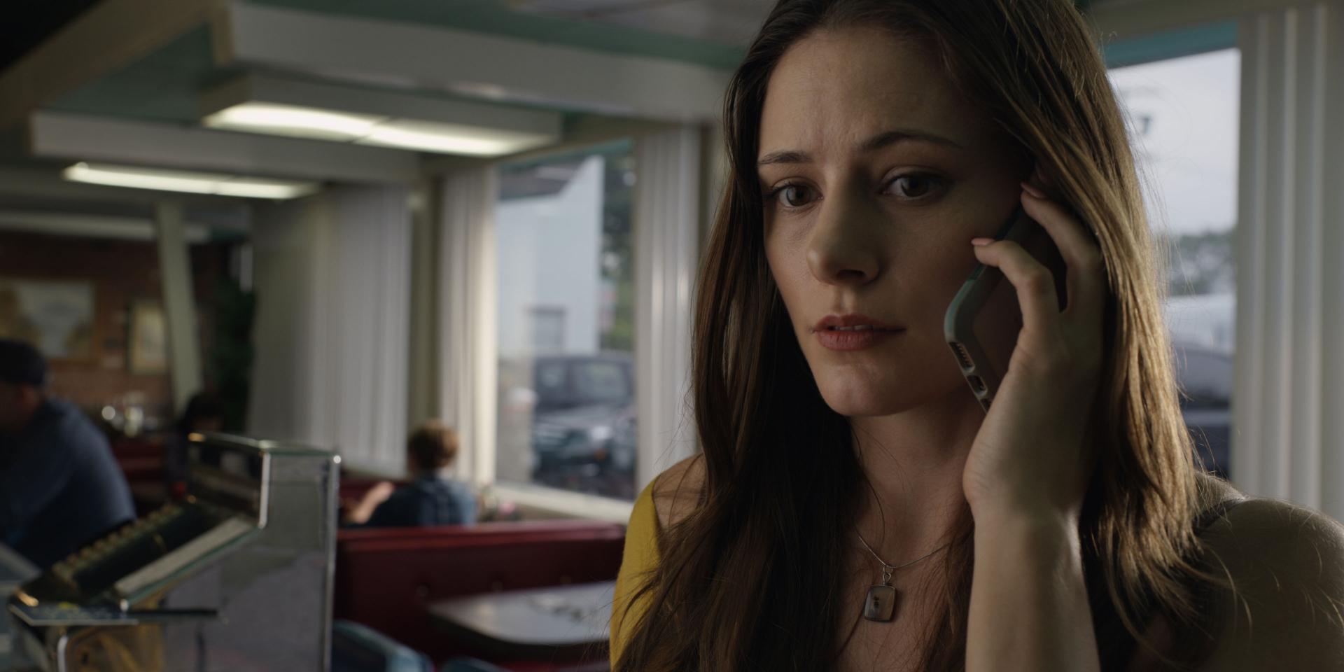 A woman is talking on a mobile phone while sitting in a retro-style diner with large windows and natural light.