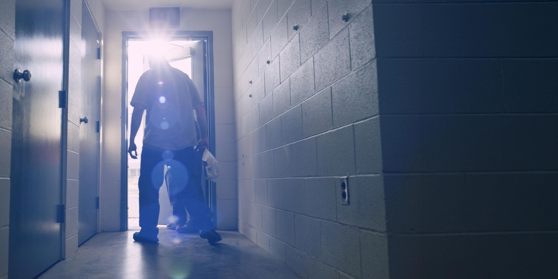 A person stands silhouetted in a brightly lit doorway at the end of a hallway with white brick walls, captured with noticeable lens flare, showcasing ZEISS optical quality.