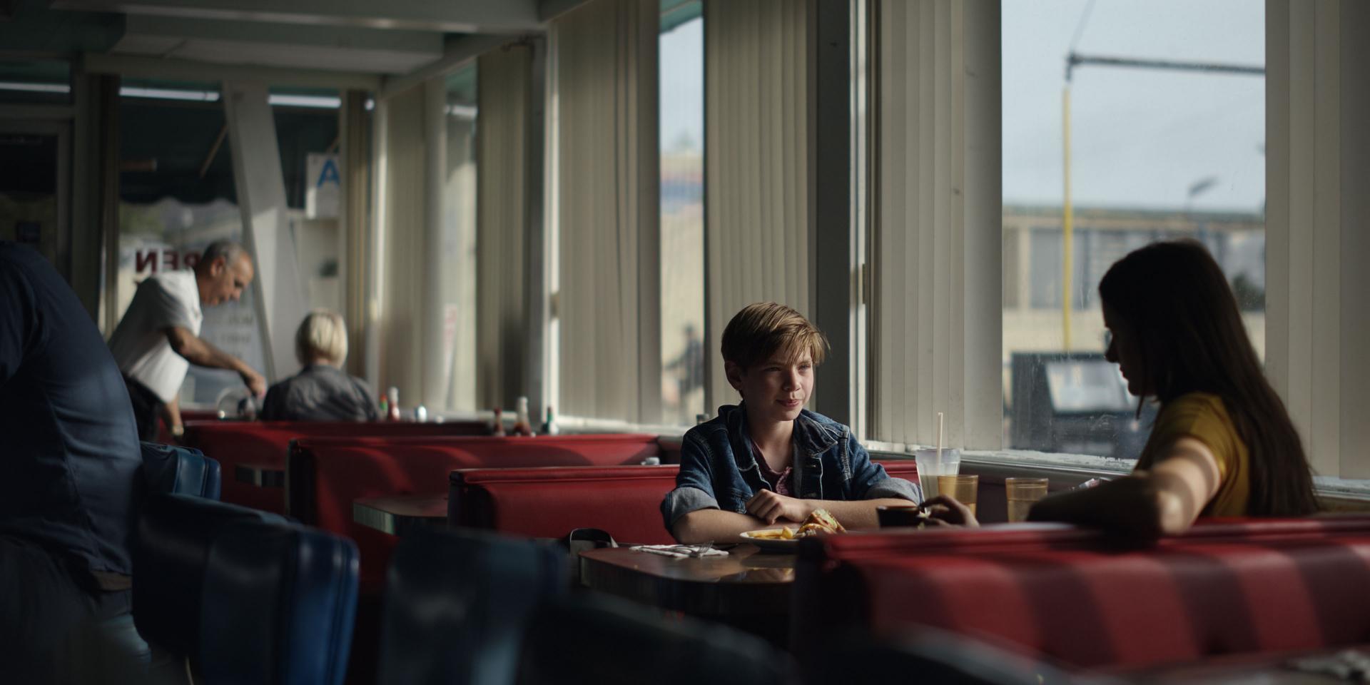 Two people sit across from each other at a table in a retro-style diner with large windows and red booth seating, captured with cinematic clarity.