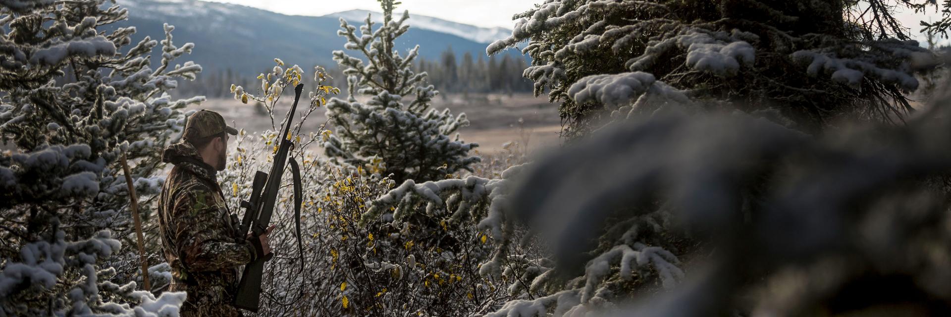 A hunter in camouflage gear holds a rifle while standing among snow-dusted pine trees in a mountainous landscape.