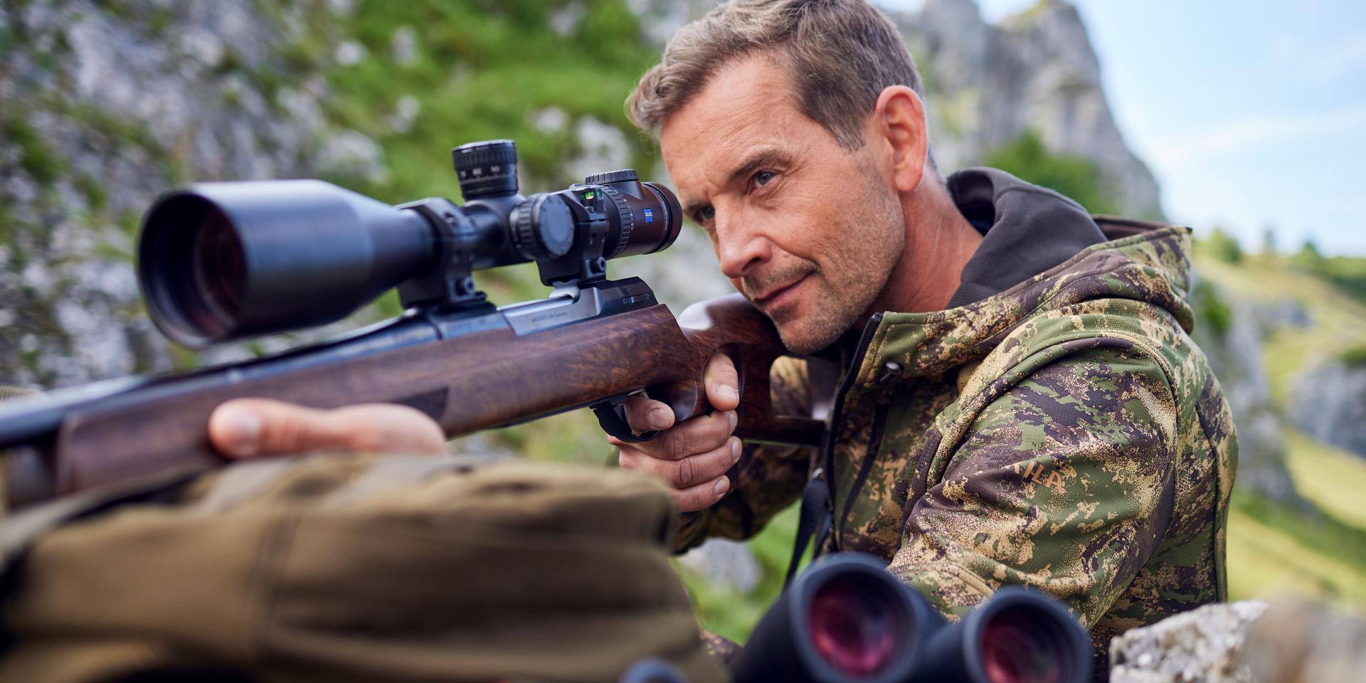 A man in camouflage clothing aims a rifle equipped with a ZEISS scope in a mountainous outdoor setting.