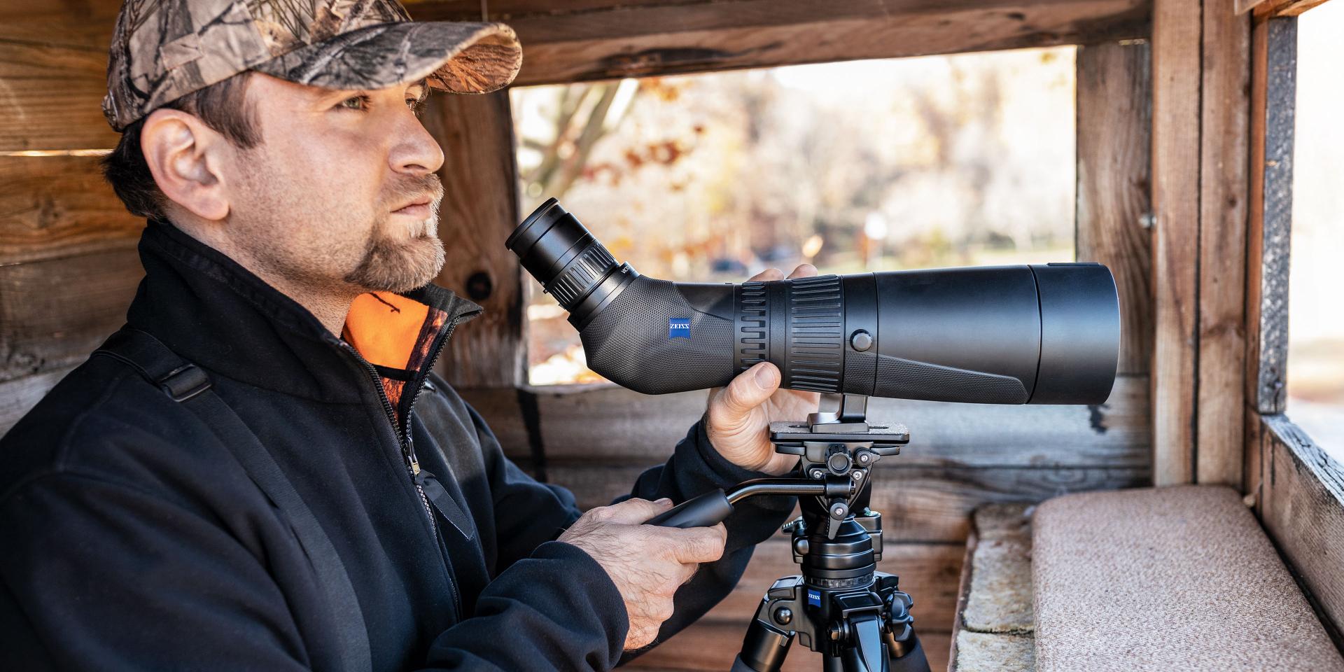 A man in outdoor gear uses a ZEISS spotting scope mounted on a tripod inside a wooden observation shelter.