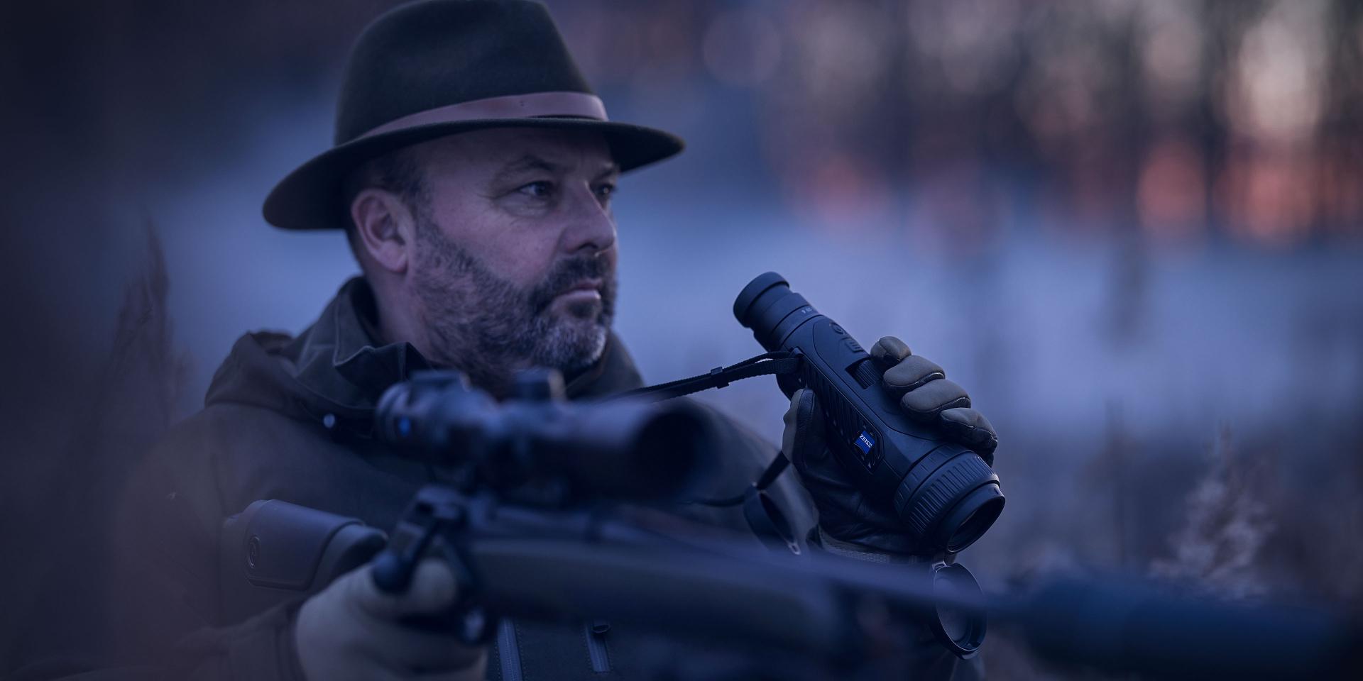 A man in outdoor clothing holds a rifle and a ZEISS thermal imaging monocular while observing his surroundings in a forest at dusk.