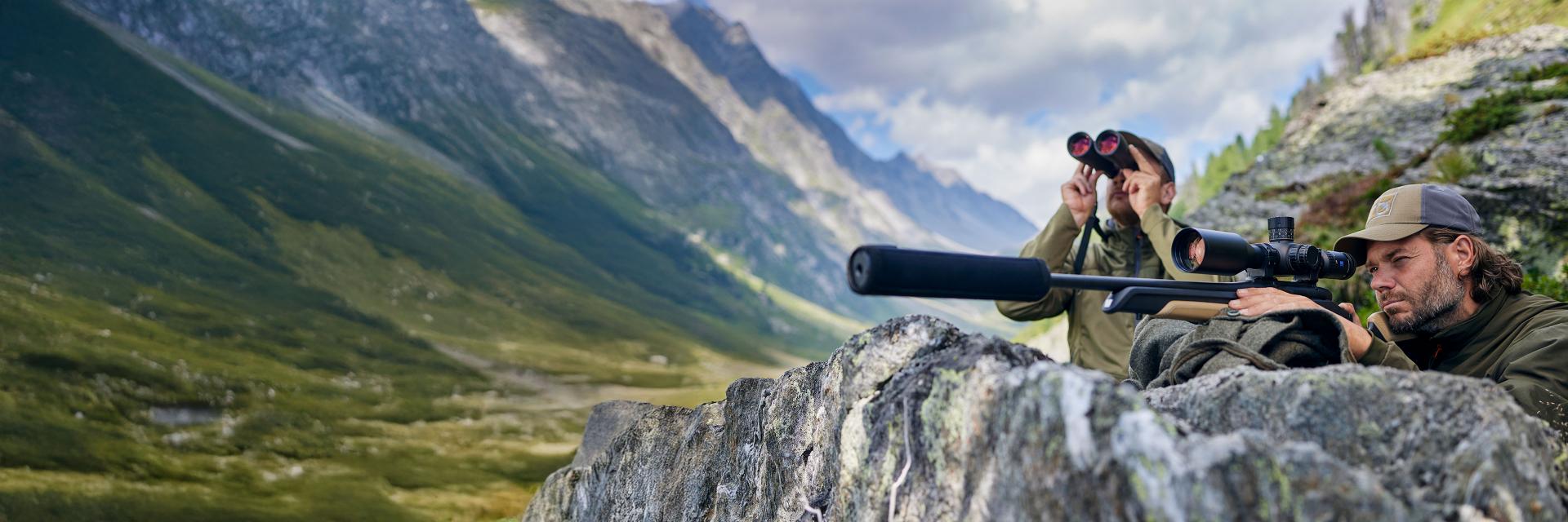 Two people in outdoor gear are positioned behind a rock in a mountainous landscape, one using ZEISS binoculars and the other aiming a rifle with a ZEISS riflescope.