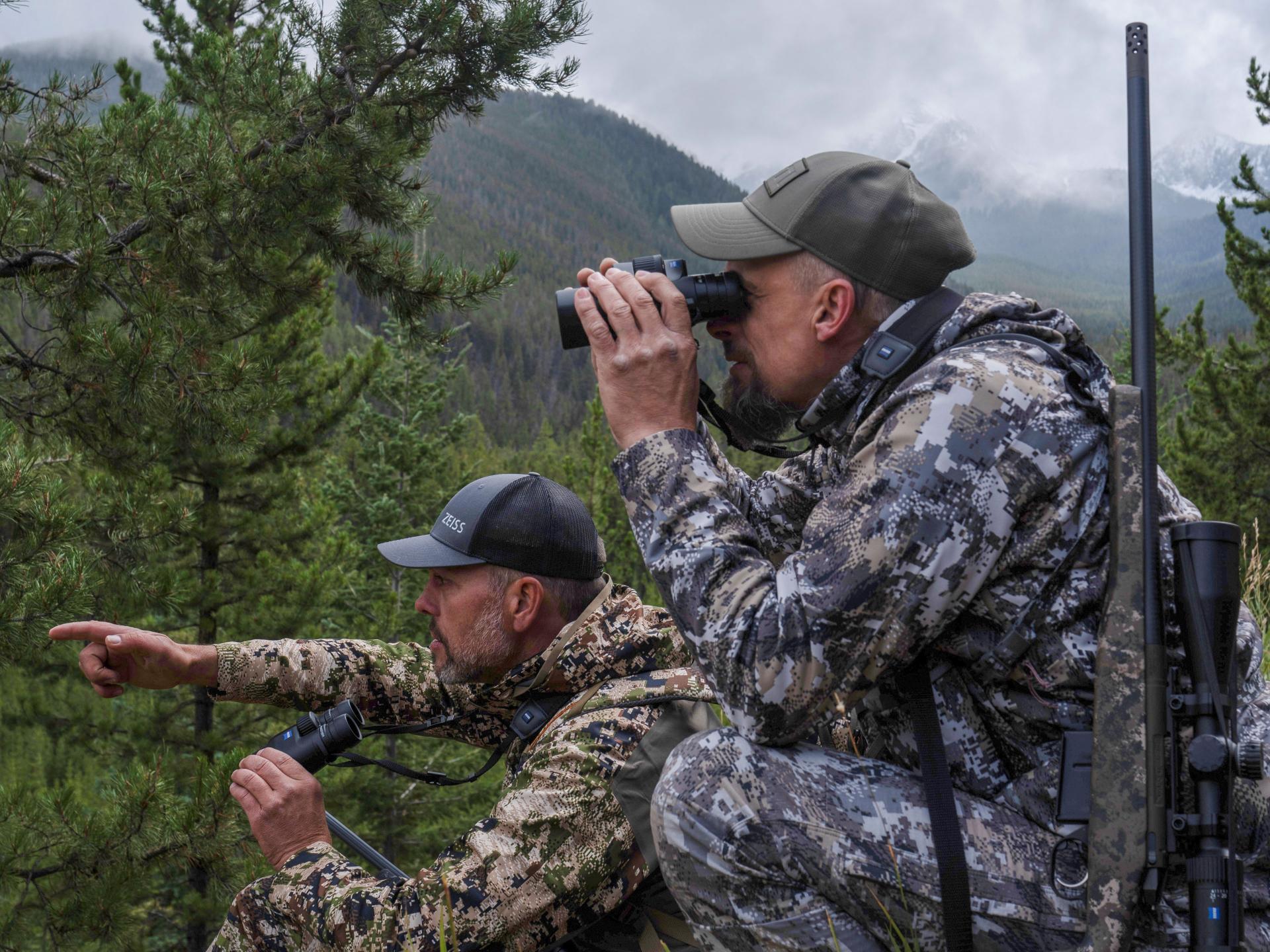 Two men in camouflage gear are observing a forested mountain landscape using ZEISS binoculars.