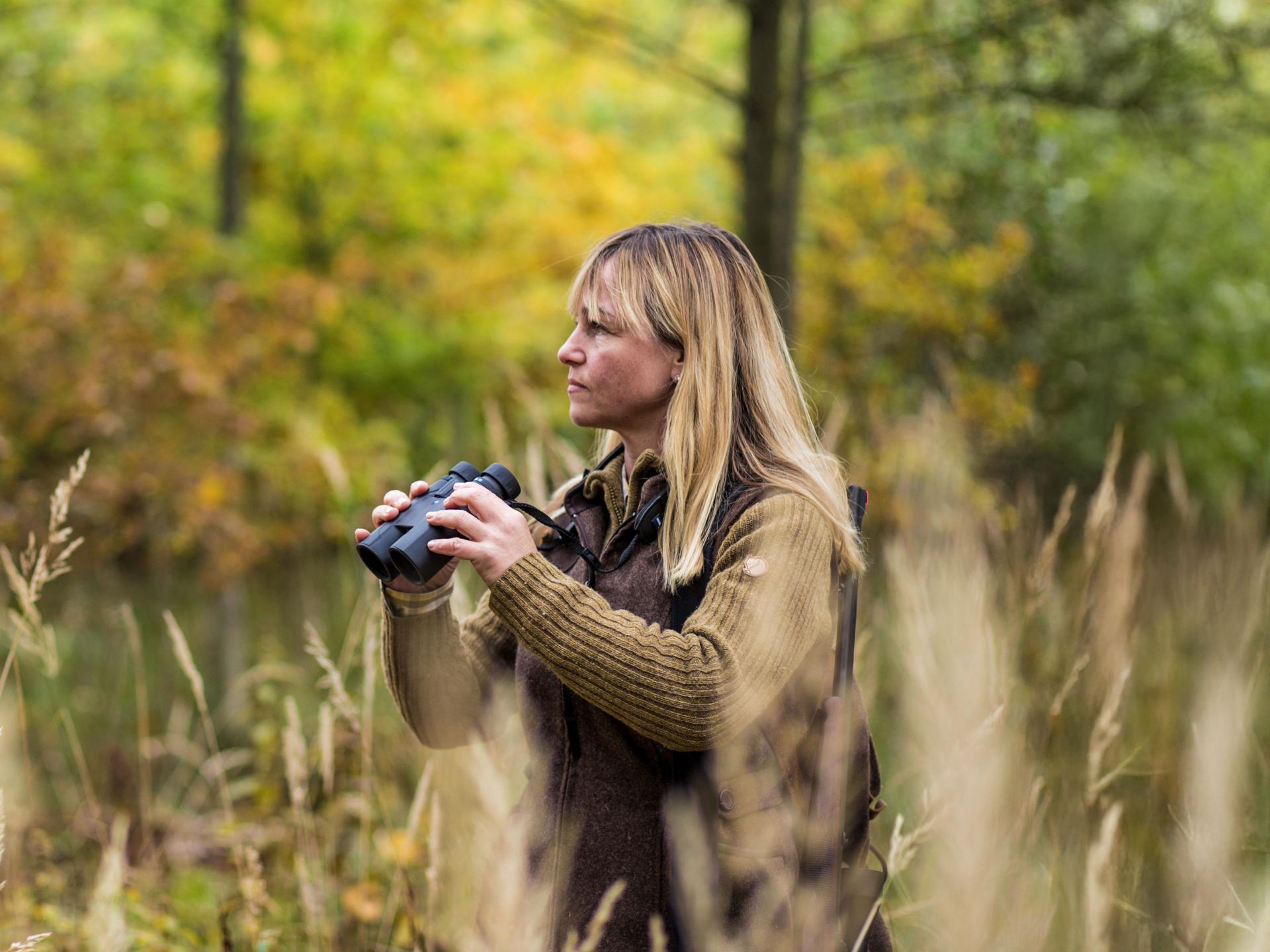 A woman stands in tall grass in a forested area, holding ZEISS binoculars and looking into the distance.