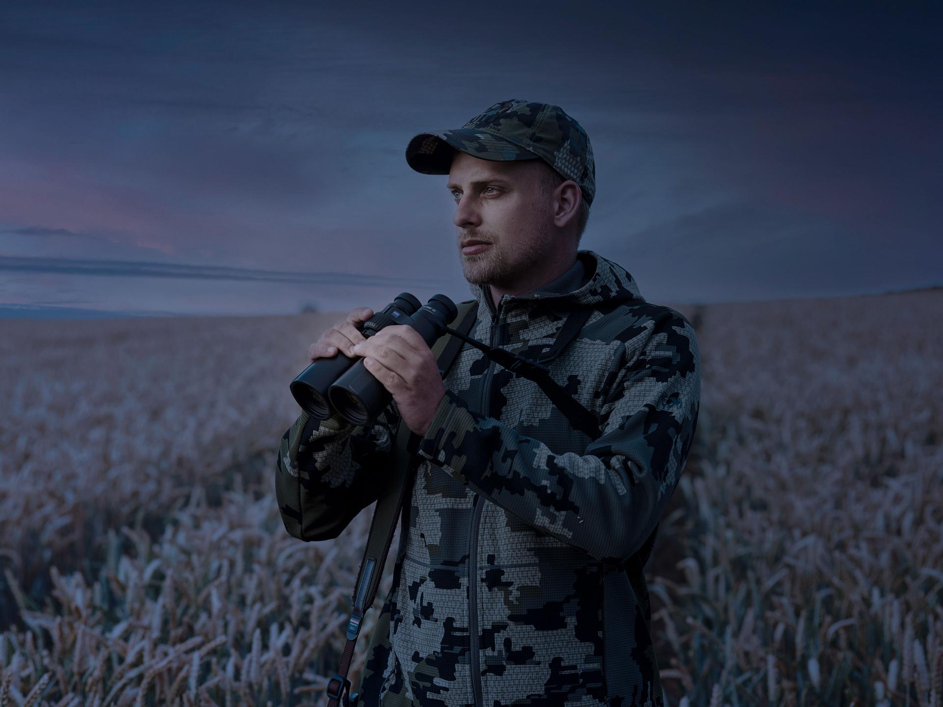 A man in camouflage clothing stands in a frosty field at dusk, holding a pair of ZEISS binoculars.