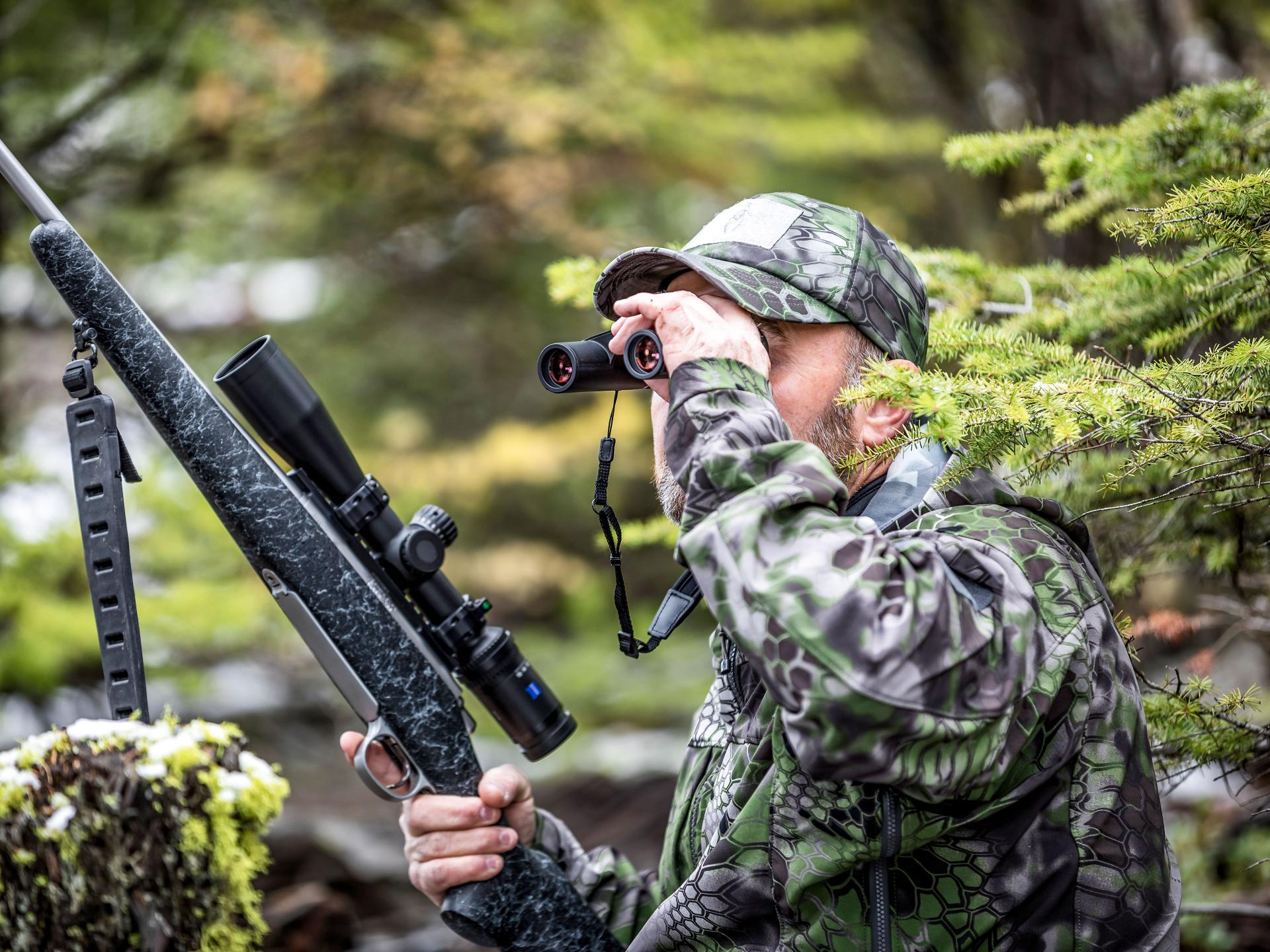 Hunter in camouflage gear holding a rifle with a mounted ZEISS scope and looking through ZEISS binoculars in a forest setting.