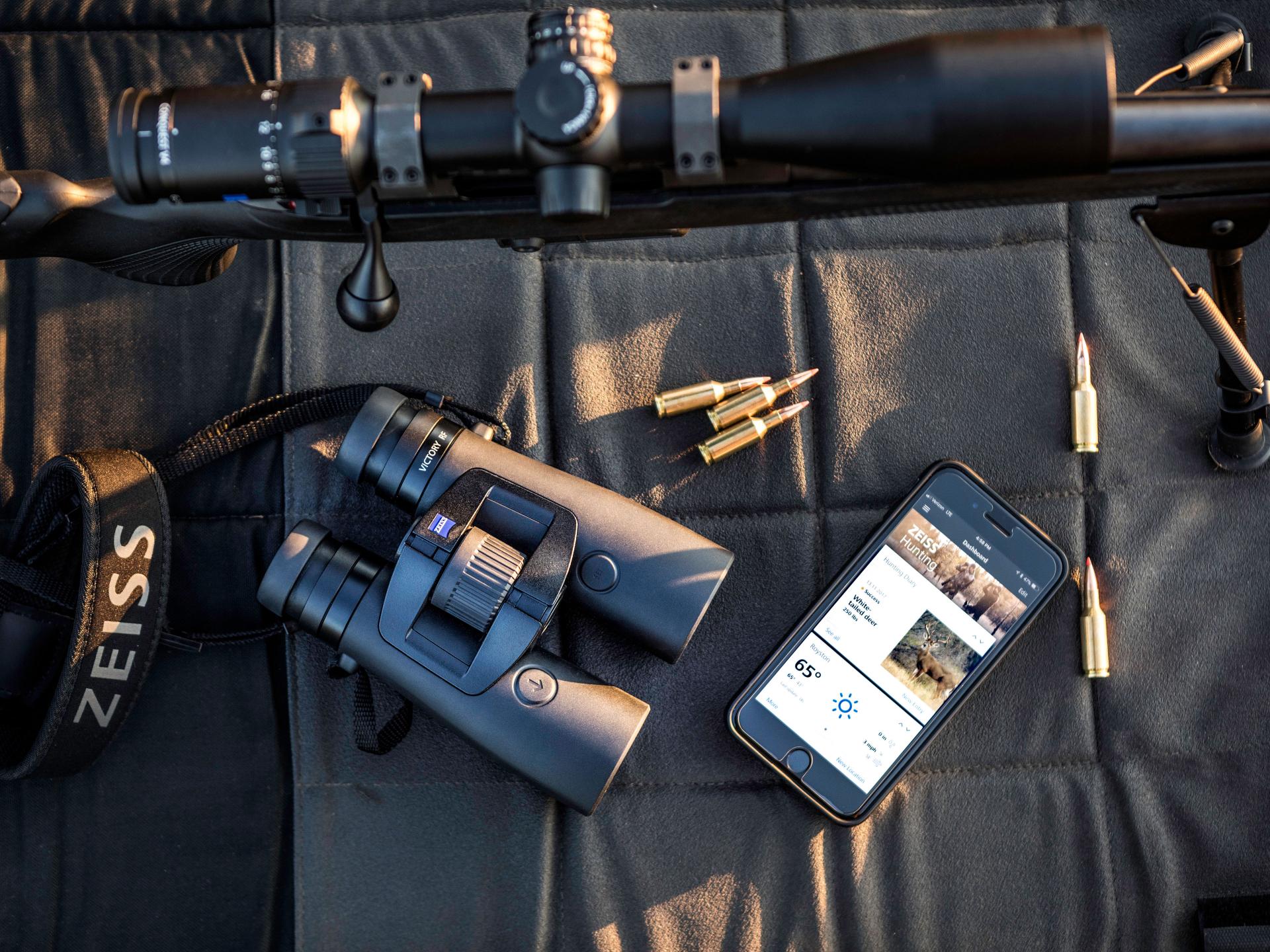 ZEISS binoculars, a ZEISS rifle scope, rifle cartridges, and a smartphone displaying a hunting app are arranged on a padded surface.