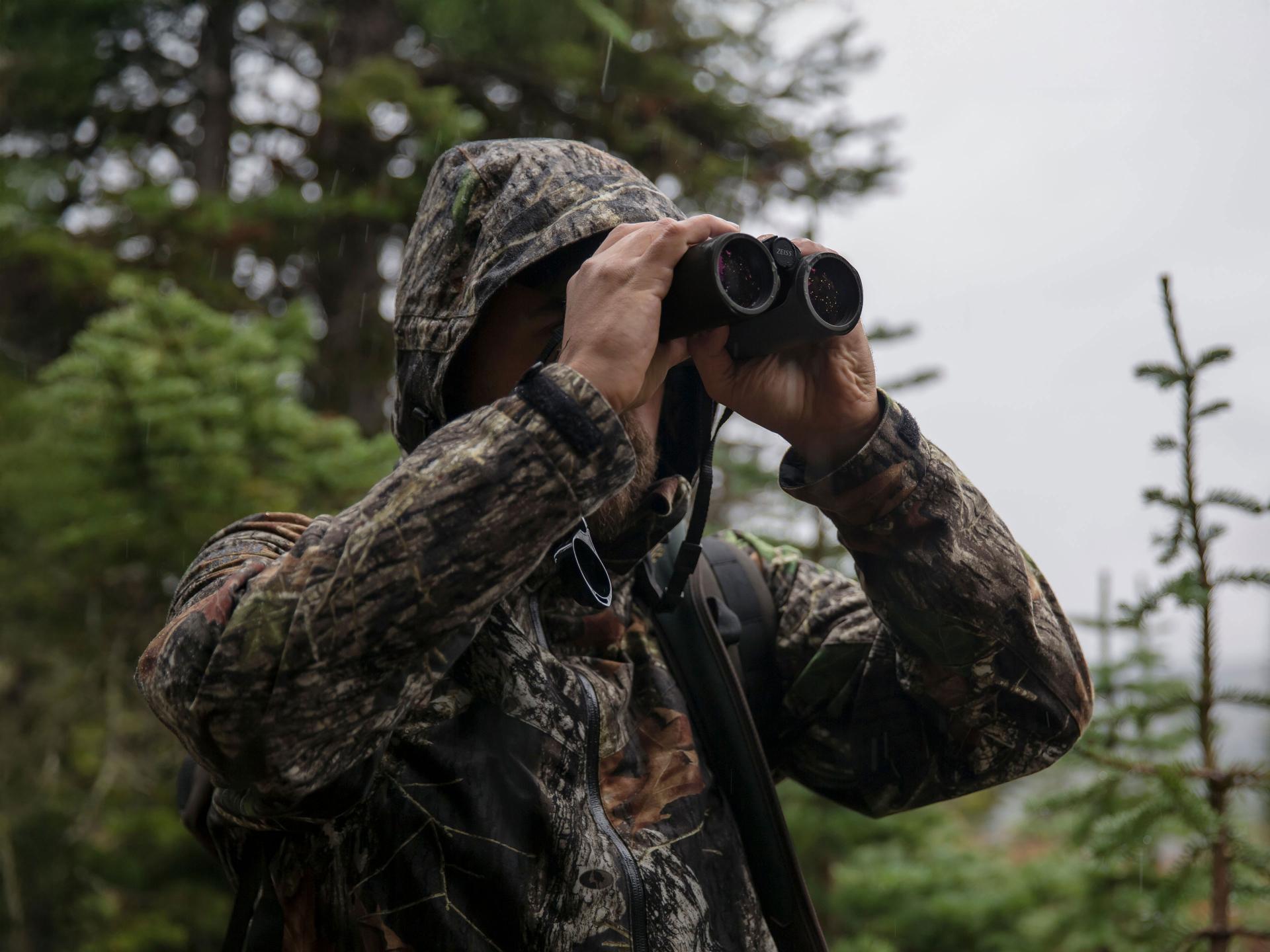 A person in camouflage clothing observes the surroundings through ZEISS binoculars in a forested area.