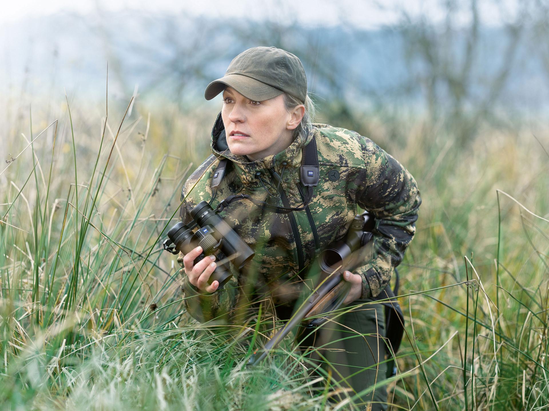 A person in camouflage clothing crouches in tall grass holding a pair of ZEISS binoculars and a rifle.