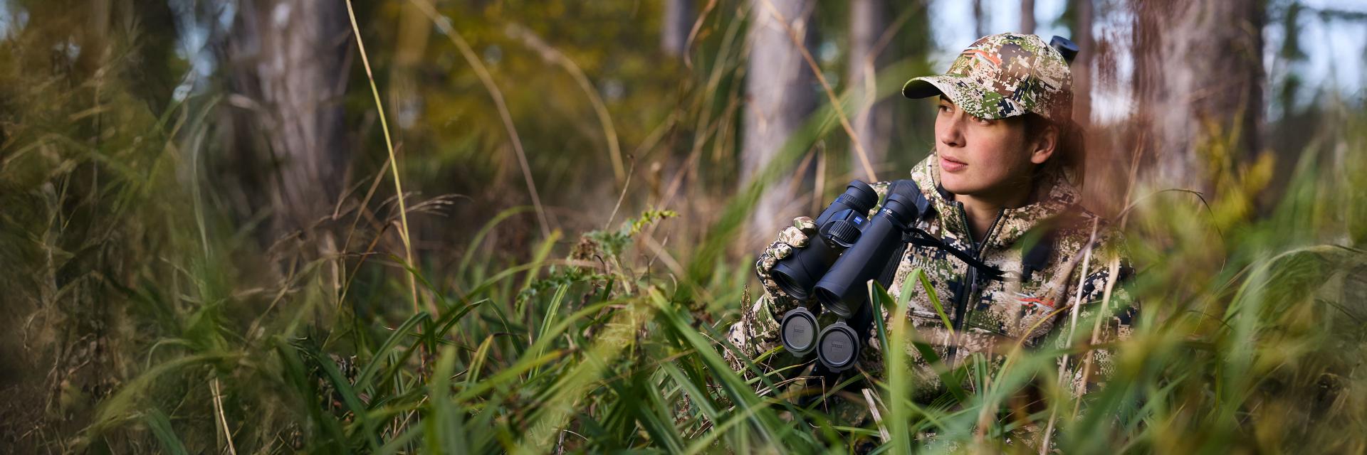 A person in camouflage clothing is sitting in tall grass in a forest, holding a pair of ZEISS binoculars.