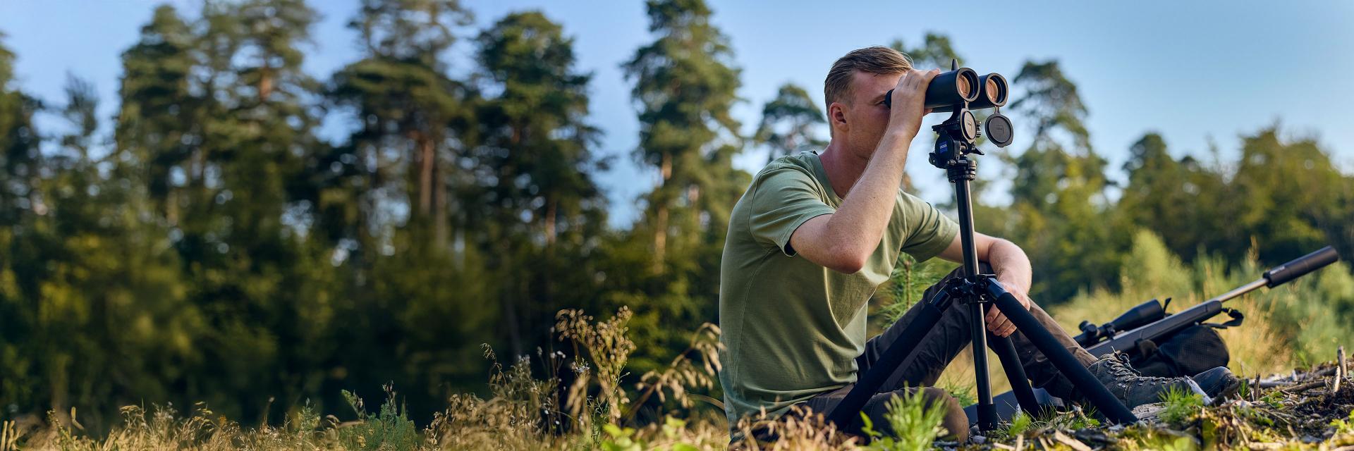 A man sits in a grassy clearing using ZEISS binoculars mounted on a tripod, with a forest in the background and a rifle resting beside him.
