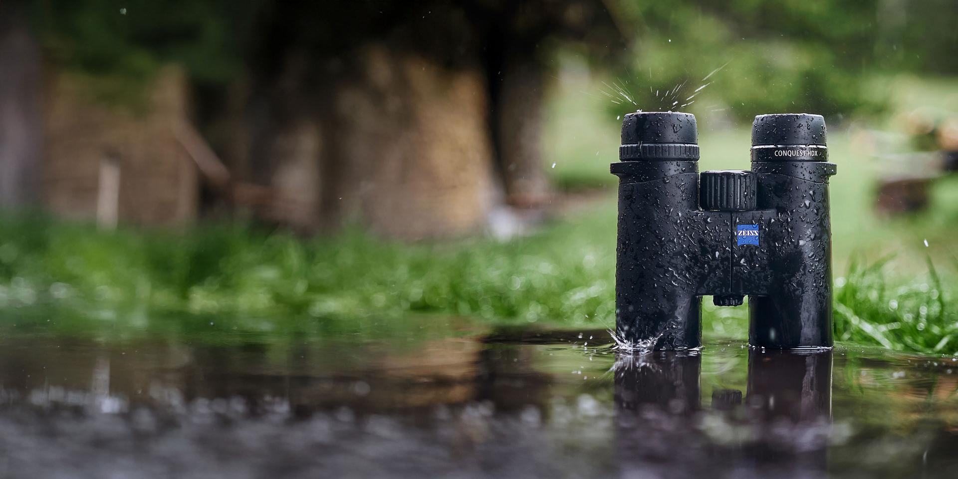 A pair of black ZEISS binoculars stands upright in a rain puddle with water droplets splashing around, set against a blurred outdoor background.