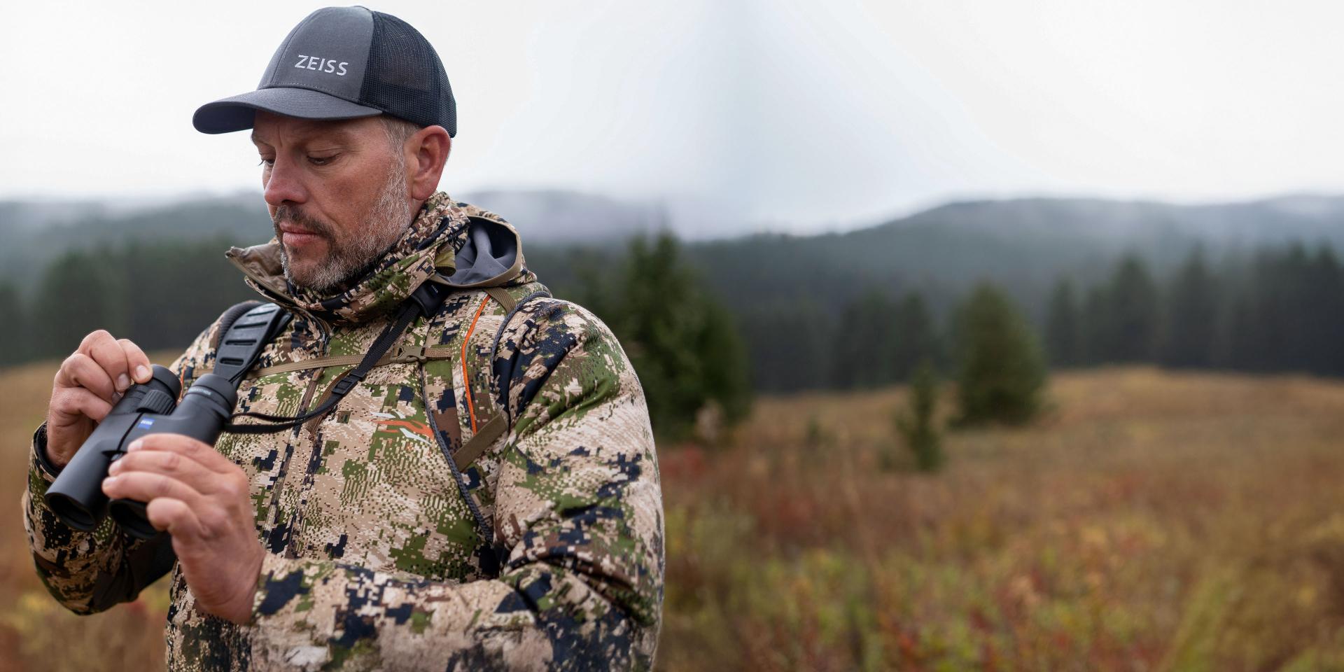 A man in camouflage gear stands in a grassy, forested landscape while adjusting ZEISS binoculars and wearing a ZEISS-branded cap.