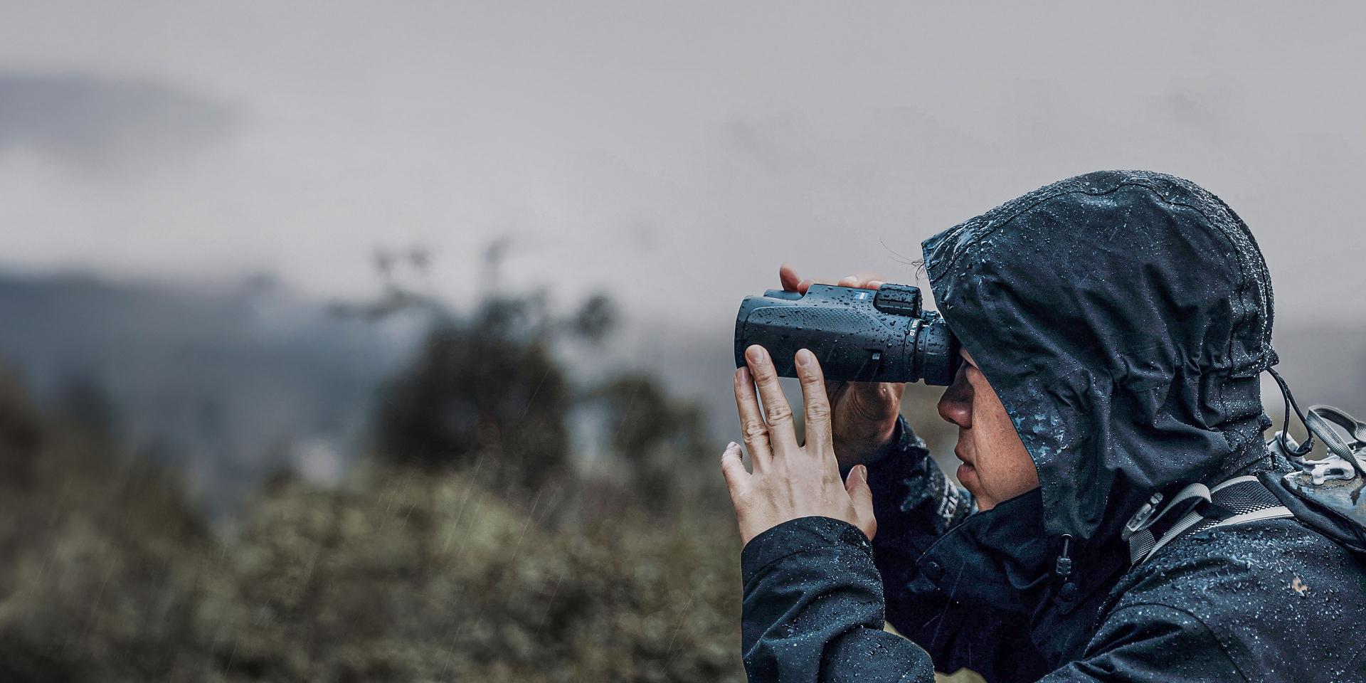 Person in a rain jacket using ZEISS binoculars to observe a landscape in rainy weather.