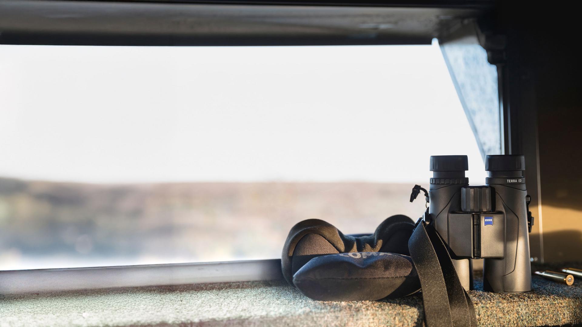 A pair of ZEISS binoculars and a neck cushion rest on a windowsill inside a vehicle with an outdoor view.