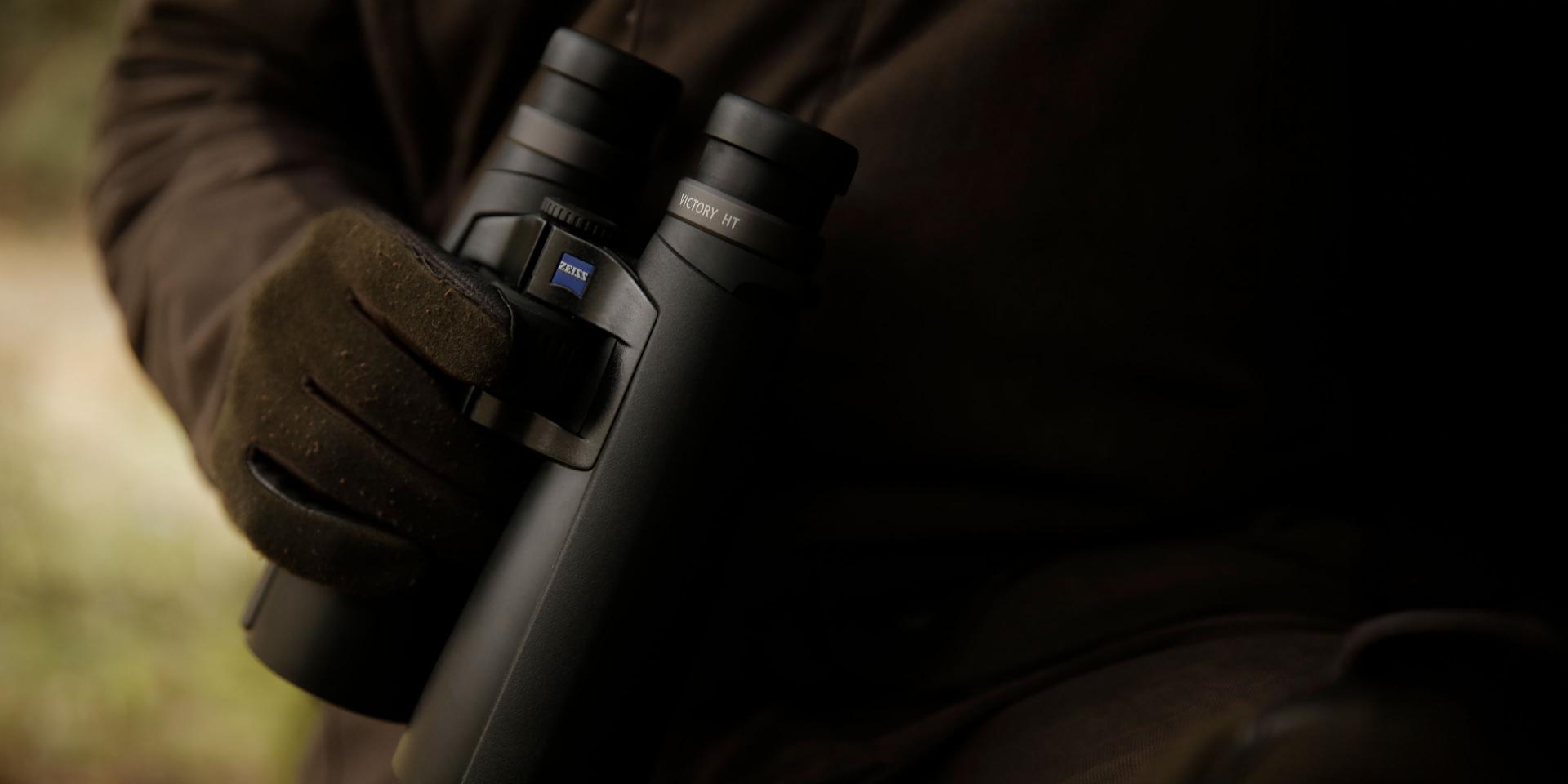 A person wearing a glove is holding a pair of black ZEISS binoculars in an outdoor setting.