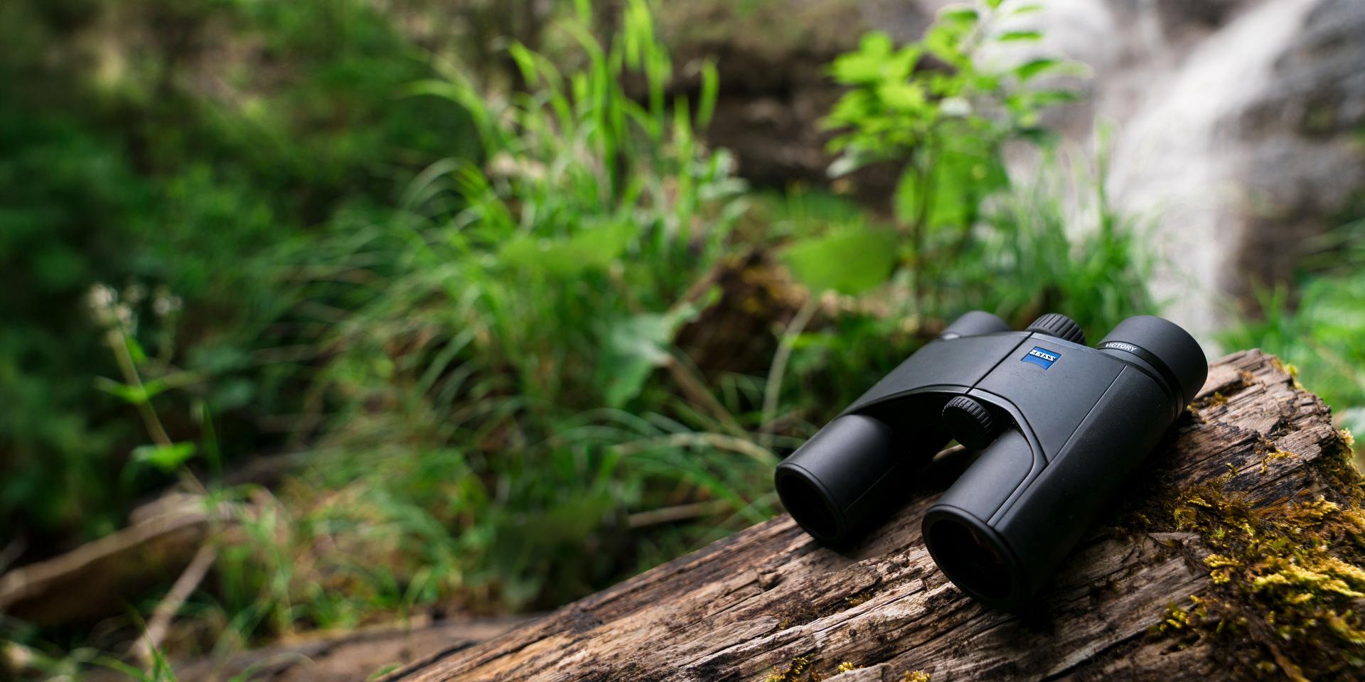 A pair of ZEISS binoculars rests on a mossy log in a lush forest near a waterfall.