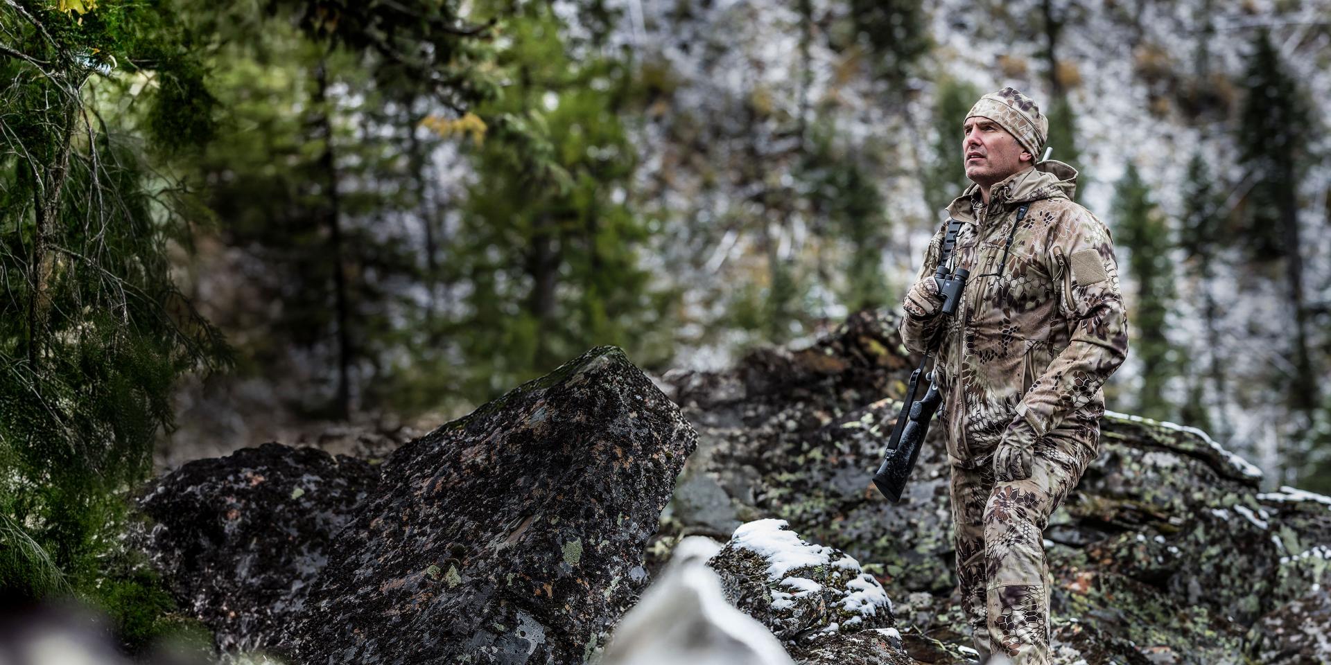 A hunter in full camouflage gear stands in a mountainous forest landscape, holding a rifle equipped with a ZEISS scope.